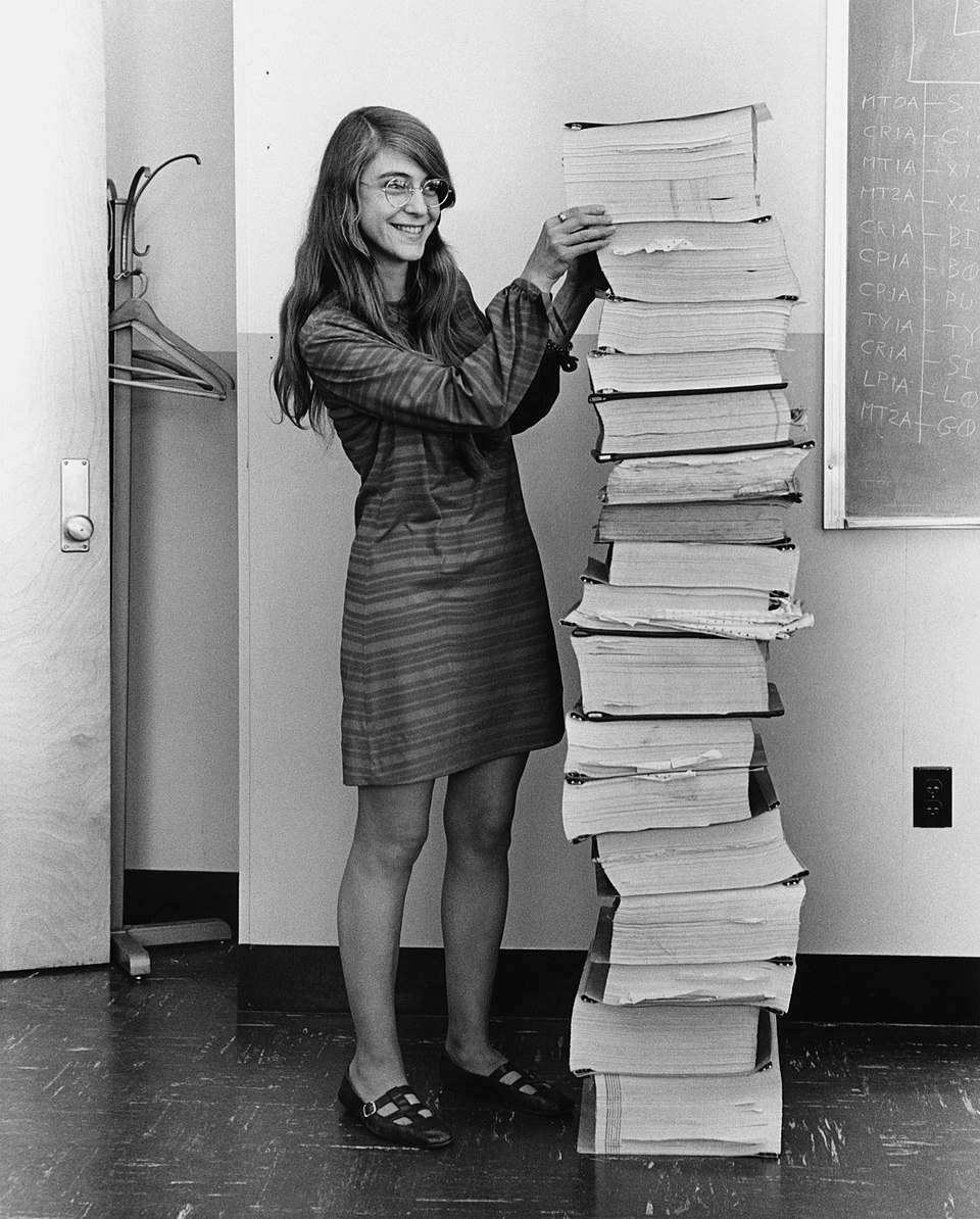Margaret Hamilton standing beside a stack of printed Apollo Guidance Computer source listings nearly as tall as she is